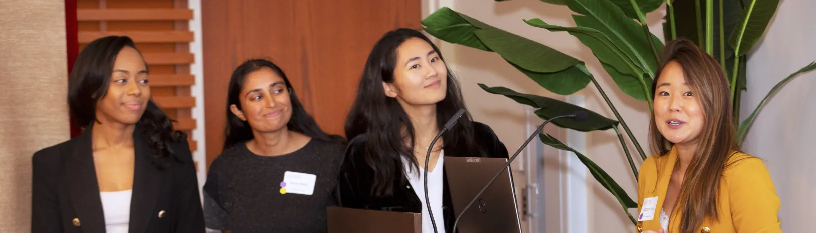 Student speaks at Stanford GSB Impact Fund event with diverse, professionally-dressed students listening.