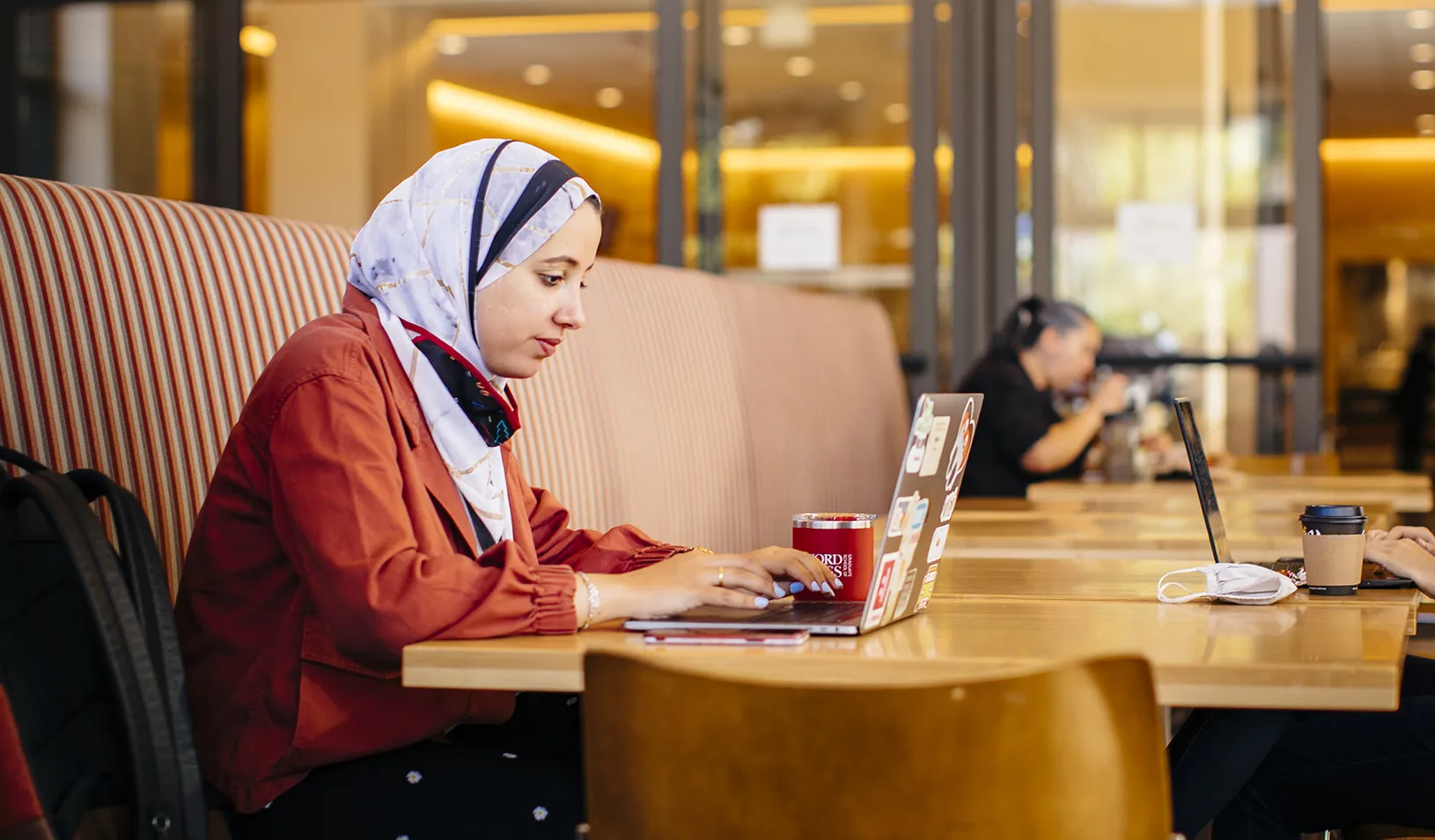 A female student in a headscarf sitting in Arbuckle Dining Hall on Stanford GSB campus, working on her laptop. Photo by Elena Zhukova