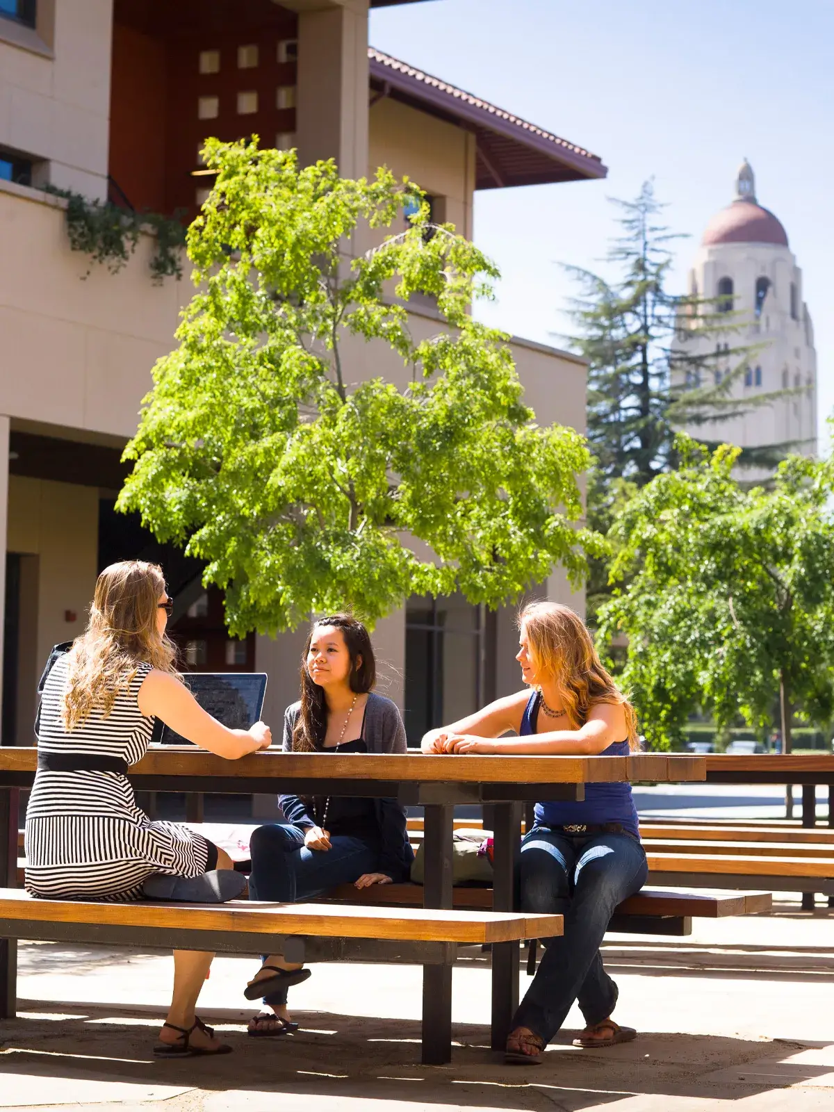 a group of students in conversation at a picnic table outside at the GSB, with Hoover tower in the background.