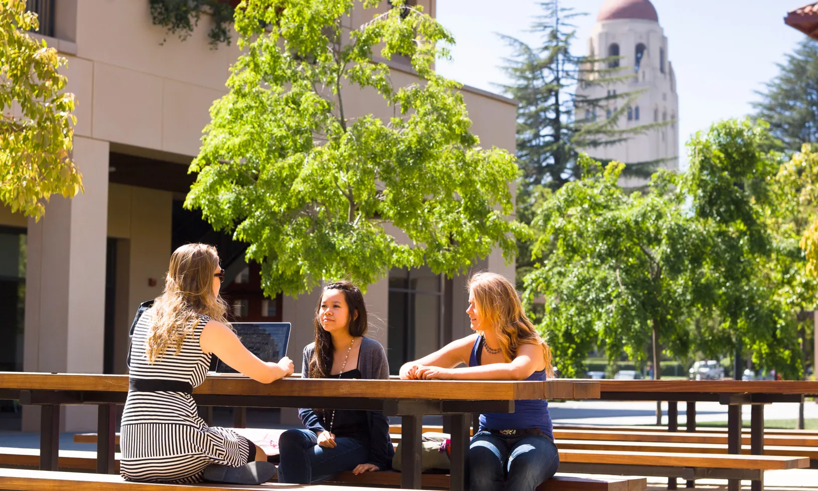 a group of students in conversation at a picnic table outside at the GSB, with Hoover tower in the background.