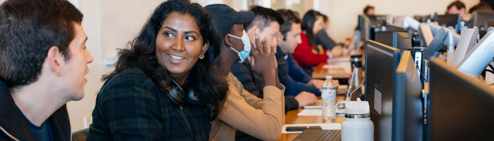 Group of students in a classroom with a woman with medium length black hair and headphones around her neck turned toward her neighbor in conversation