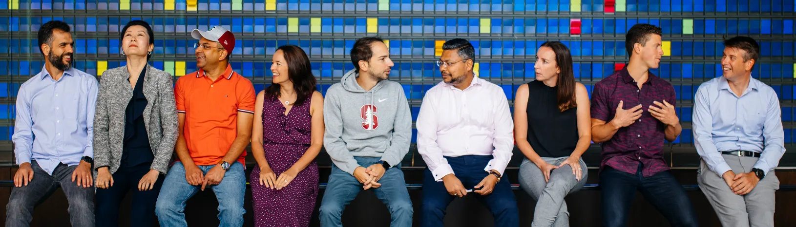 Diverse group of nine male and female students in front of color wall that is mostly blue with yellow accents