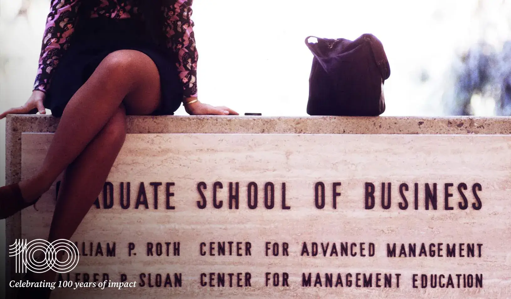 Older photo of a women sitting atop a sign for the Graduate School of Business. Credit: Courtesy of Susan Phillips, Anne Thornton, and Barbara West, with special thanks to Luther Nussbaum