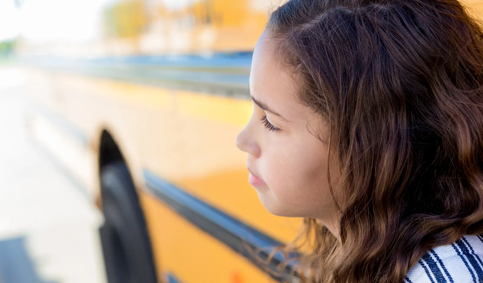 Mixed race schoolgirl daydreams while standing in front of a school bus. Credit: iStock/SDI Productions