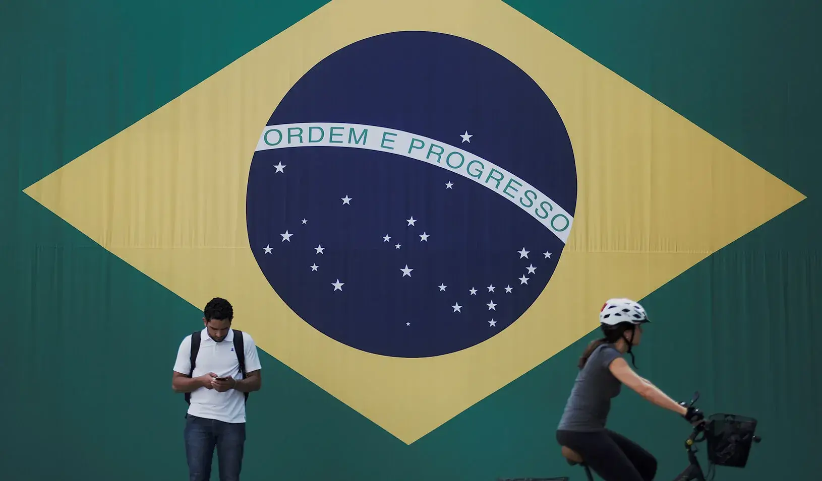 A man checks his mobile phone as a woman riding a bike passes next to a big Brazilian flag in Sao Paulo, Brazil. Credit: Reuters/Nacho Doce