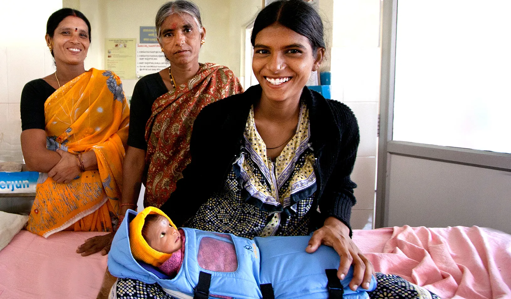 Three women near a baby wrapped in an Embrace warmer