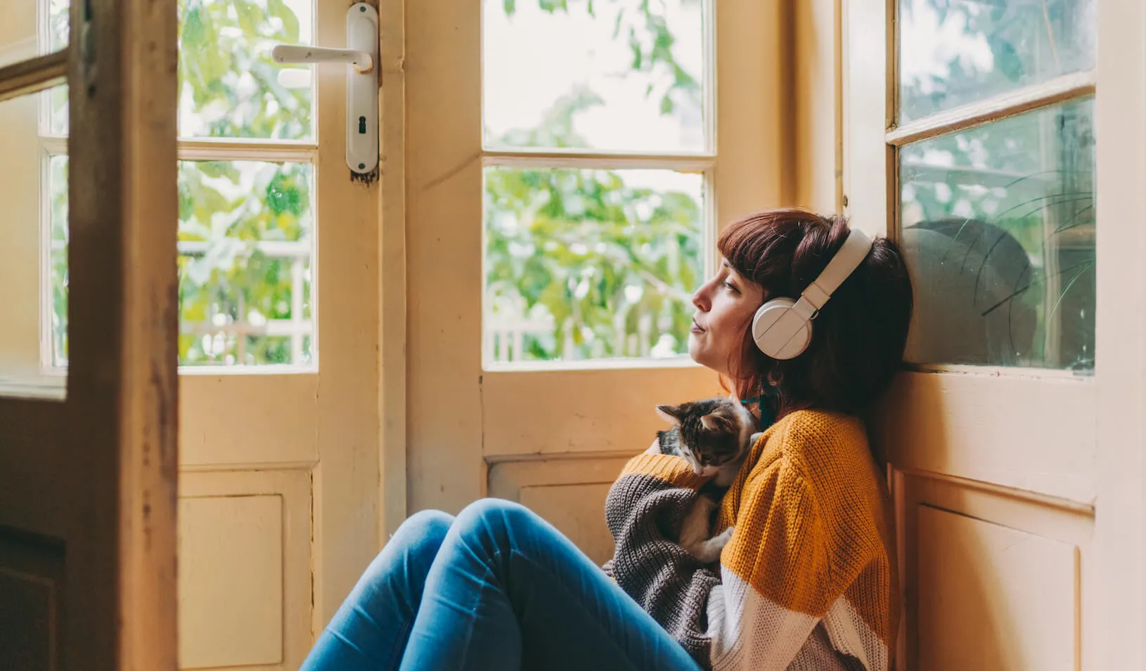 Woman cuddles cat and looks out the window while listening to a podcast in headphones. Credit: iStock/Martin Dimitrov