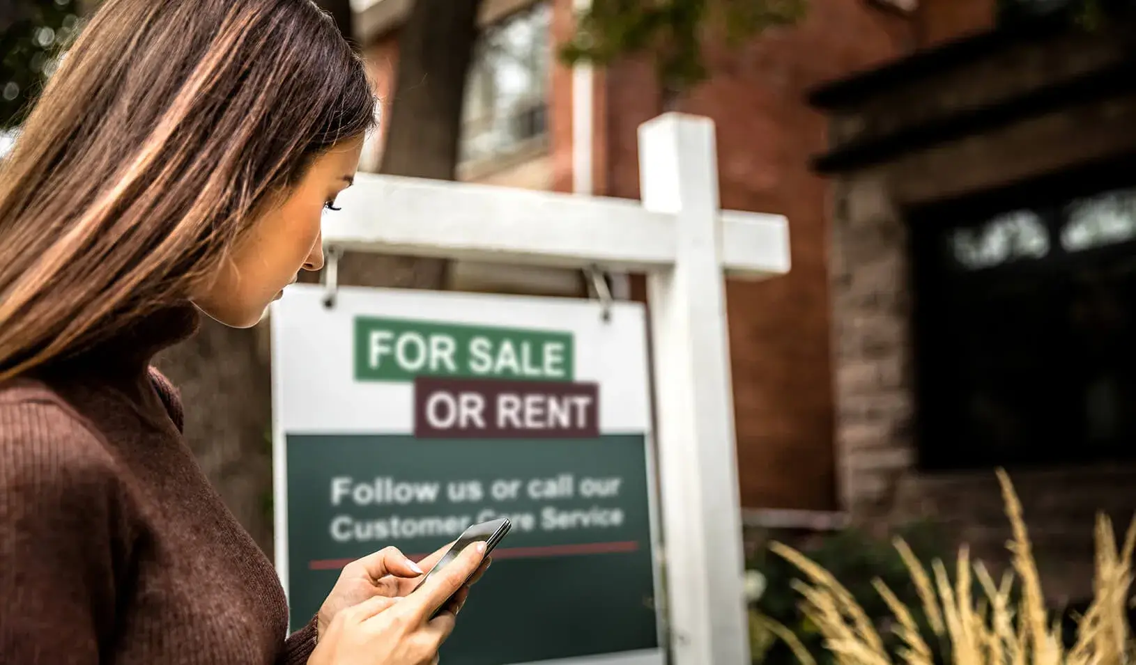 A woman taking a photo of a for-sale sign in front of a house. Credit: iStock/Leonardo Patrizi