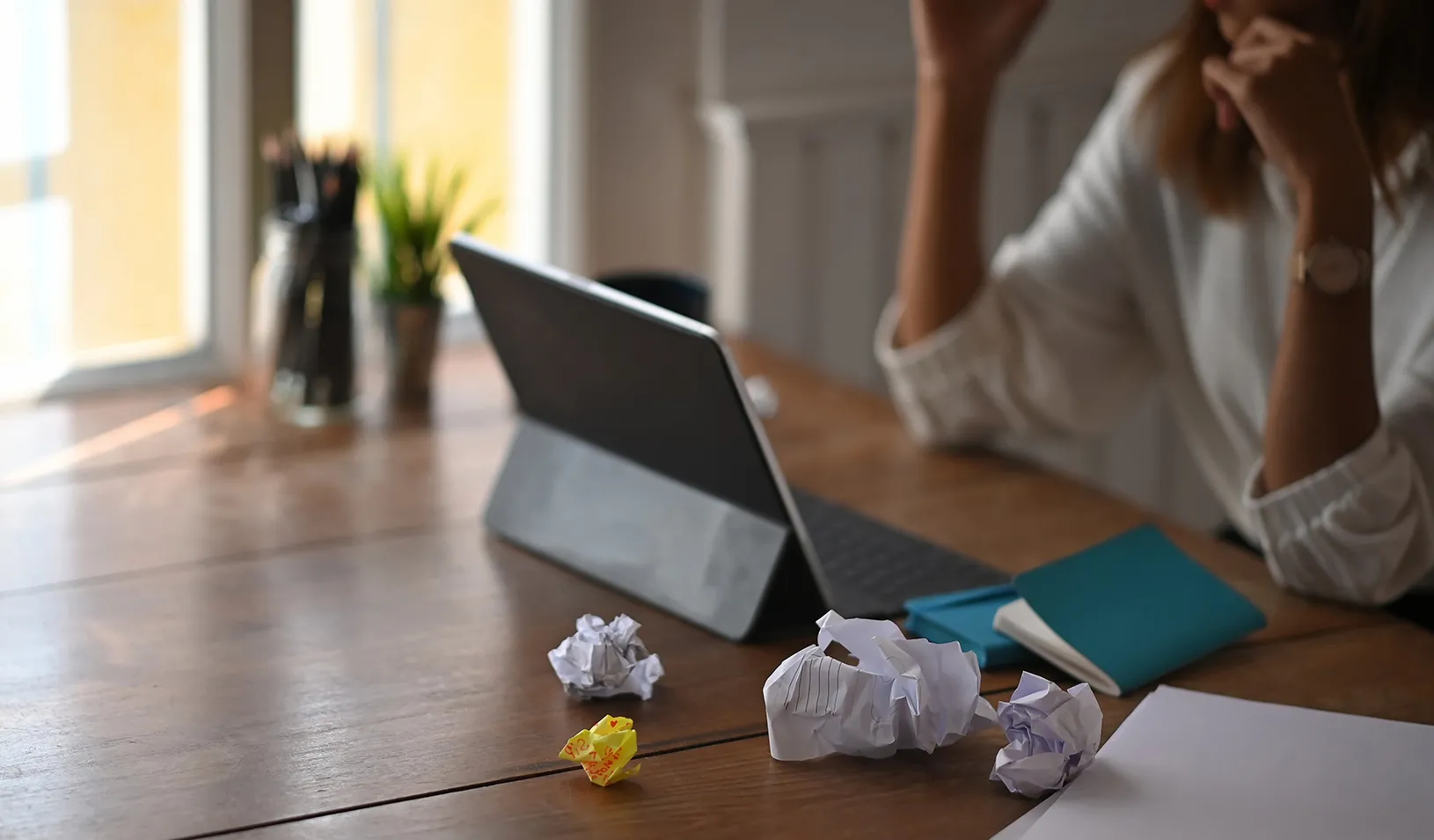 Woman sits at table looking at tablet, with crumpled papers around her, feeling stressed. Credit: iStock/PrathanChorruangsak