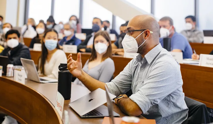 GSB students in a classroom wearing masks, watching intently as a male student holds his hand forward while speaking to the class. | Photo by Elena Zhukova.