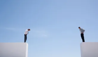 Two men standing on either side of a wide open gap on white platforms in front of a blue sky, looking down in puzzlement. iStock/Martin Barraud