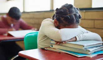 A young Black student sits at her desk with her head down on a pile of books. iStock/LumiNola