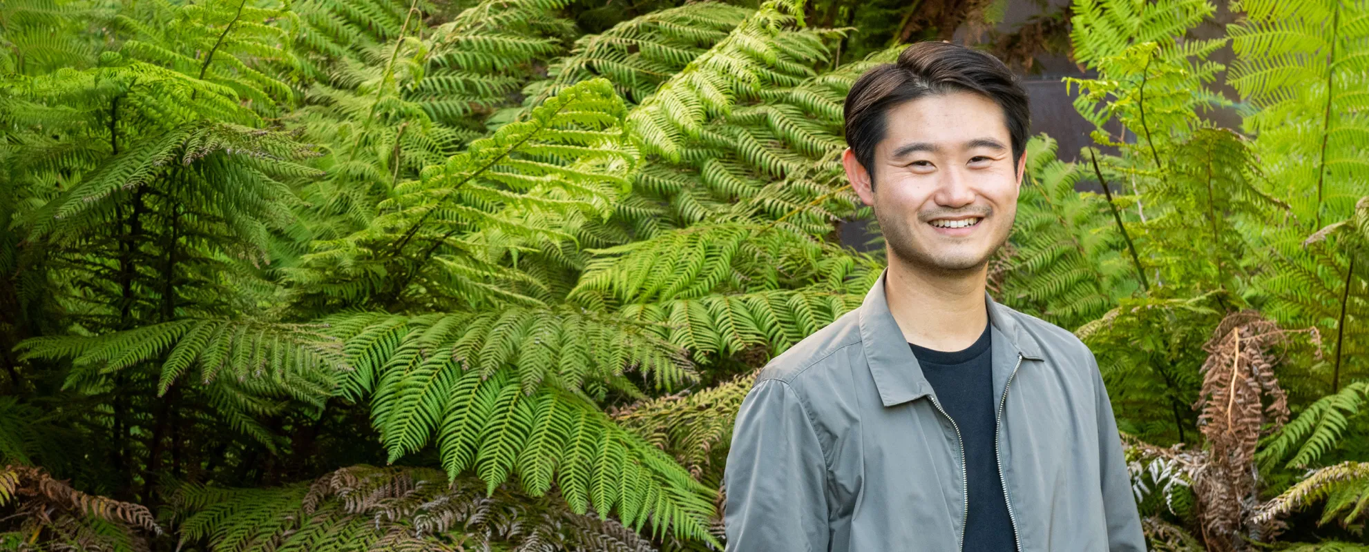Stanford GSB Professor Charles Chu standing outside in front of ferns smiling at the camera. | Credit: Kiefer Hickman.