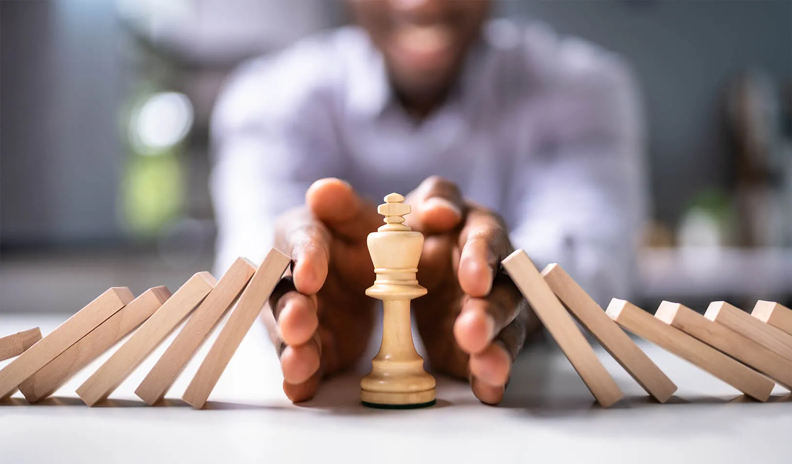 A businessman's hands protect a chess king piece from falling wooden blocks or dominoes. | Credit: iStock/AndreyPopov.