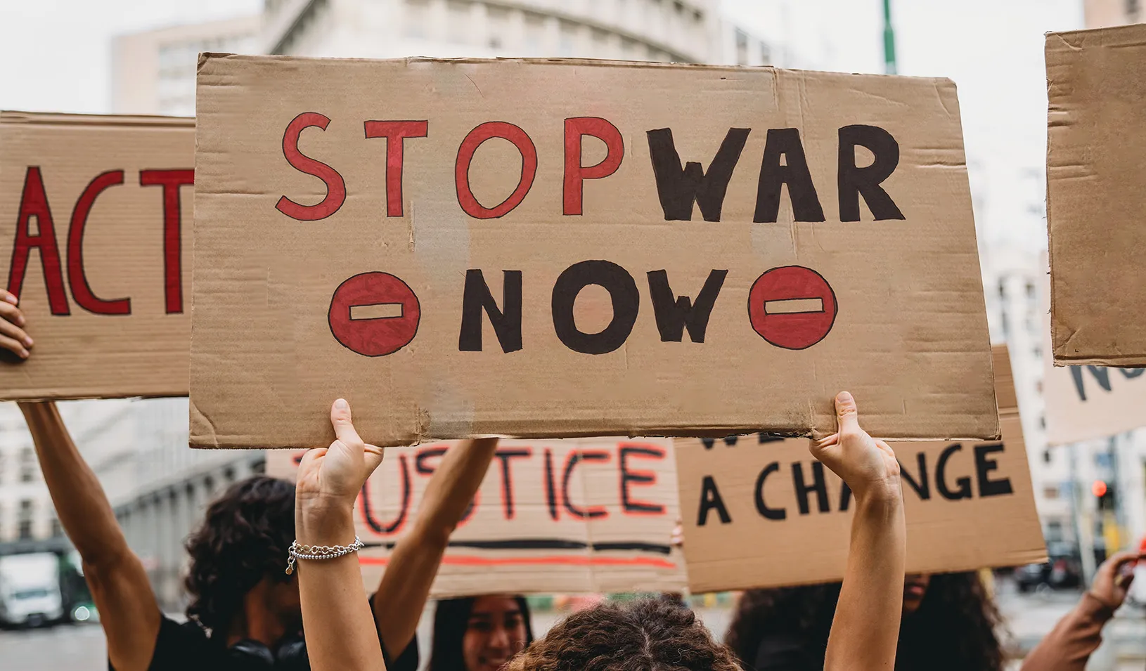 People holding protest signs on a cloudy day in an unknown city. The foremost sign says "stop war now." Photo by iStock/FilippoBacci