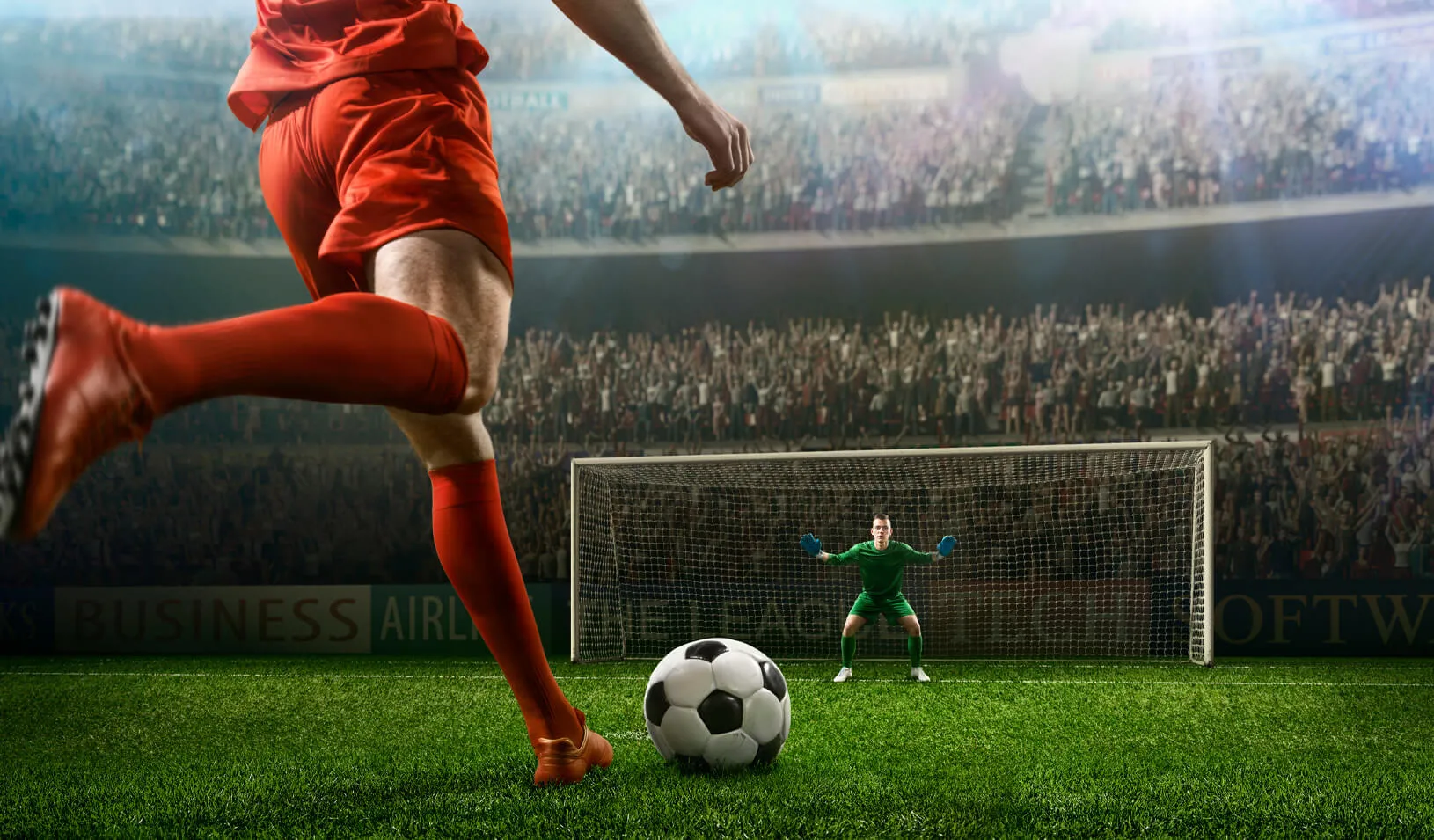 A male soccer player kicks a ball and soccer goalie jumping in motion for a ball while defending his gates on wide angle panoramic image of an outdoor soccer stadium or arena full of spectators under a sunny sky. Credit: iStock/Dmytro Aksonov