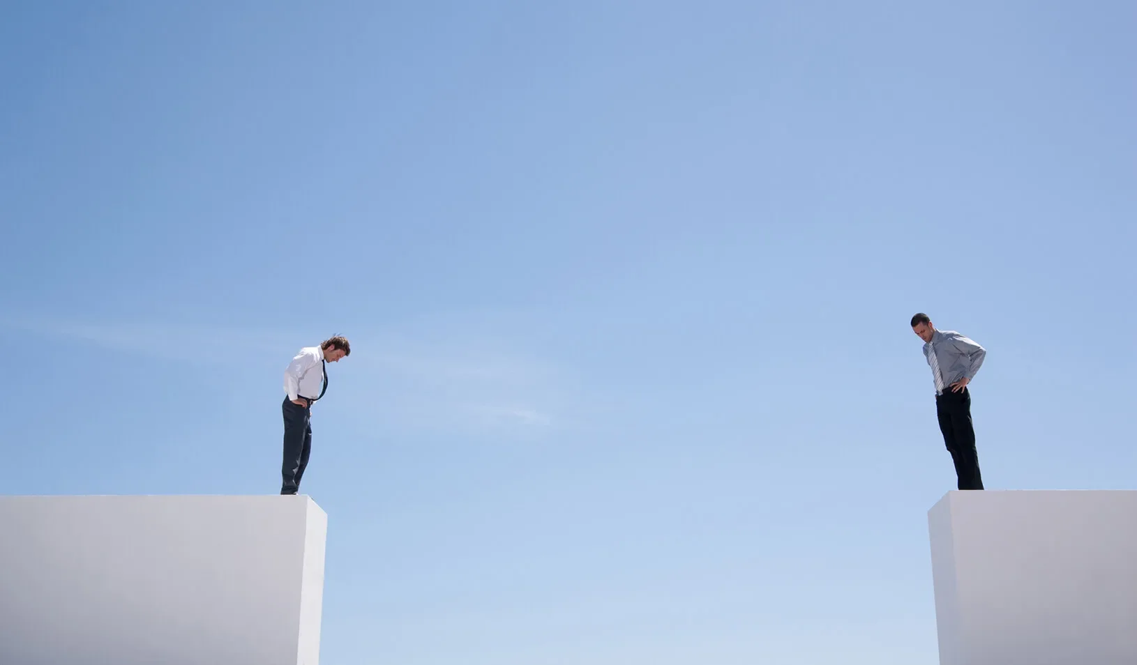 Two men standing on either side of a wide open gap on white platforms in front of a blue sky, looking down in puzzlement. iStock/Martin Barraud